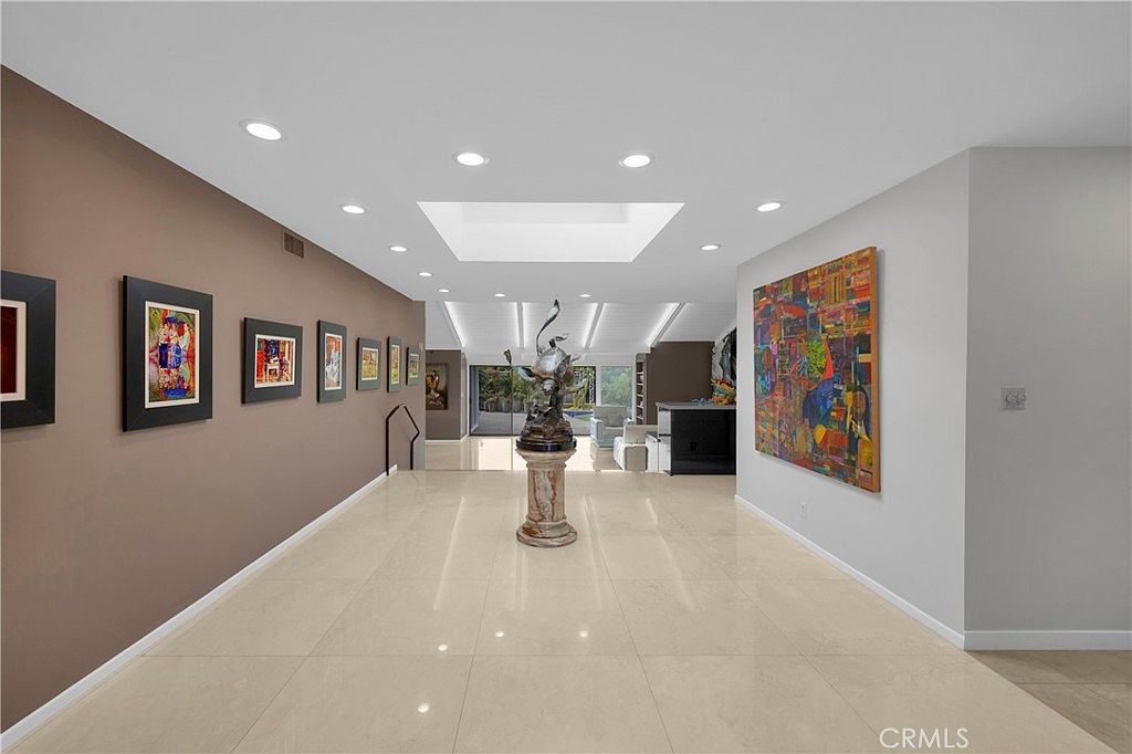 Long skylit gallery foyer with hardwood-toned floor, sculpture on a marble plinth, and a wall of collected paintings on a saddle-brown accent wall
